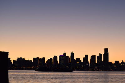 Sea by silhouette buildings against clear sky during sunset