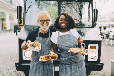 Smiling senior owner with assistant holding food plate while standing against commercial land vehicle