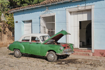 Trinidad, cuba. retro green car with open hood near house with blue walls with open door and window.