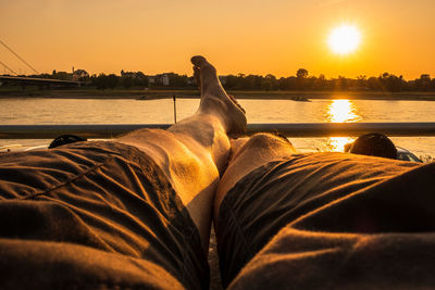 Muscular legs person in shorts relaxing on riverbank watching sunset pov