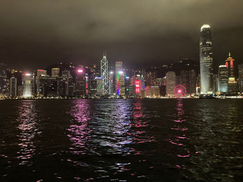 Illuminated buildings by river against sky at night