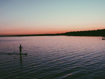 Silhouette man paddleboarding in river against clear sky during sunset