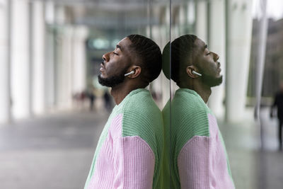 Man with eyes closed listening music through wireless in-ear headphones leaning on glass wall