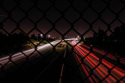 Light trail on highway against sky seen from chainlink fence