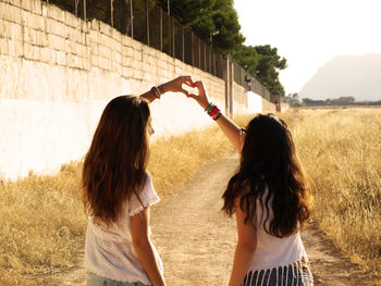 Rear view of sisters making heart shape while standing on dirt road