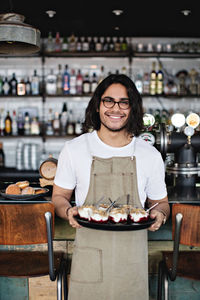 Portrait of smiling waiter holding dessert in tray while standing against counter in restaurant