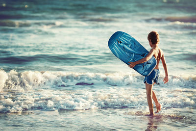 Rear view of boy carrying surfboard at seashore