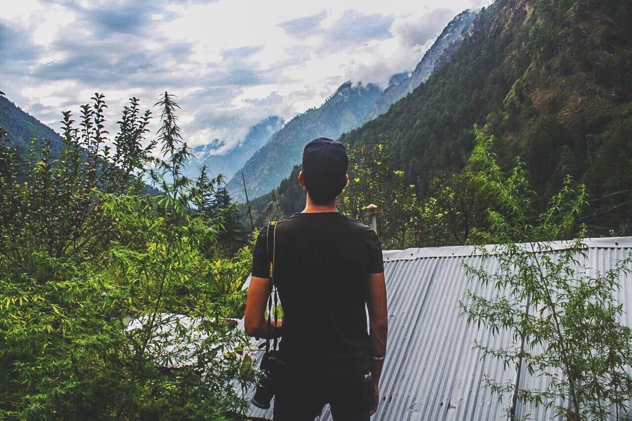 REAR VIEW OF MAN STANDING AGAINST MOUNTAIN