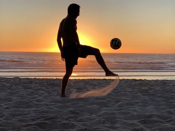Shirtless man kicking ball at beach against sky during sunset