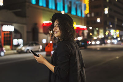 Happy woman looking away while holding smart phone in city at night