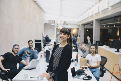 Portrait of happy female computer programmer with colleagues sitting at desk in background