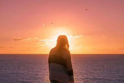Rear view of woman standing at beach against orange sky