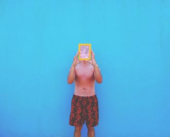 Man holding picture frame in front of face while standing against blue wall