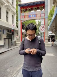 Portrait of young asian man standing on sidewalk and using smart phone against arch and buildings.