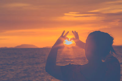 Woman making heart shape towards sun while standing by sea during sunset