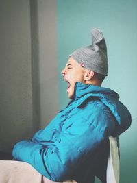 Side view of man yawning while sitting against blue wall