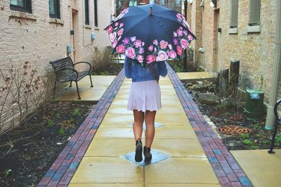 Rear view of young woman walking under umbrella on footpath amidst houses