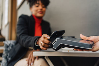 Crop african american female client putting cellphone on terminal in hand of waitress and paying for order in cafe