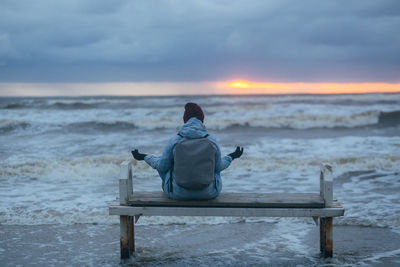 Rear view of woman sitting at beach against sky during sunset