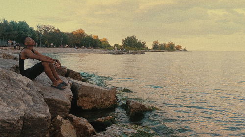 Man sitting on rocks at sea shore against sky