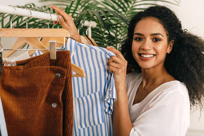 Portrait of smiling young woman holding clothing while standing by rack at home