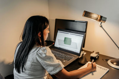 Rear view of young businesswoman sitting at desk, using laptop and 3d design software
