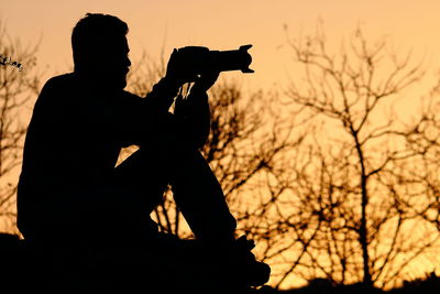 Silhouette man photographing while sitting by bare tree against orange sky