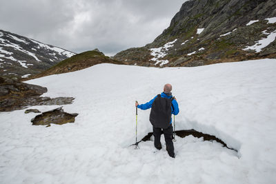 Rear view of hiker walking on snow covered field by rocky mountains against sky