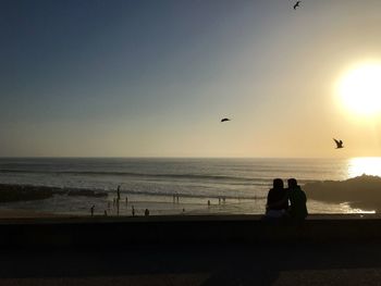 Silhouette people sitting on beach against sky during sunset