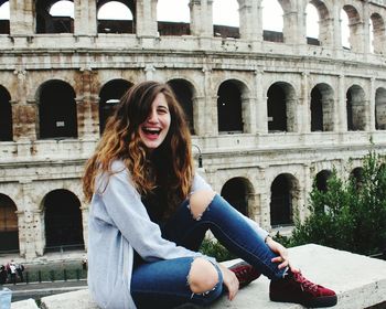 Portrait of happy woman sitting on bench against coliseum