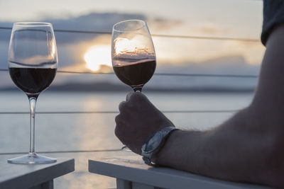 Cropped hand of man holding wine glass by sea against sky during sunset