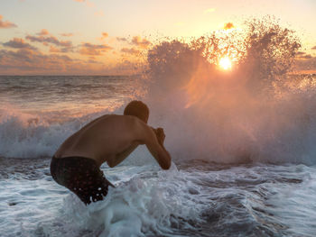 Rear view of shirtless man photographing huge wave at beach during sunset