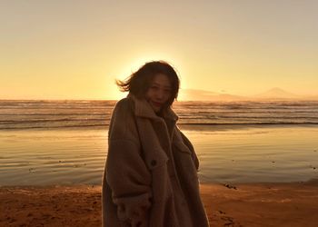 Young woman standing at beach against sky during sunset