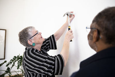 Smiling woman hammering nail on wall while standing by senior man at home