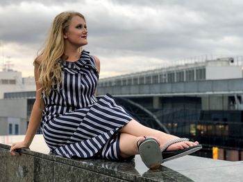 Full length of young woman sitting on retaining wall in city