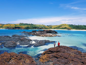 Rear view of two people standing on rock formation by sea against blue sky