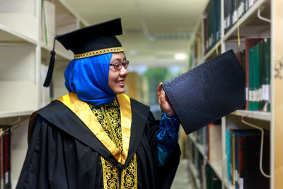 Young woman wearing graduation gown putting book on shelf in library