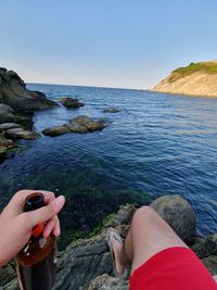 Low section of man holding beer bottle while sitting by sea