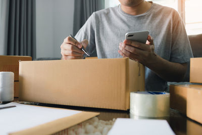 Midsection of man writing on cardboard box while holding mobile phone at home