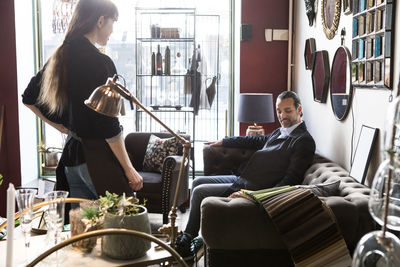 Mature male customer sitting on sofa while looking at fabric swatch with female owner standing in store