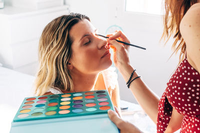 Unrecognizable female makeup artist applying stylish eyeshadows on client with eyes closed in bright beauty salon