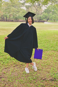 Portrait of young woman in graduation gown holding file while standing at park