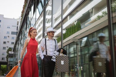 Smiling man and woman holding shopping bags while walking by building in city