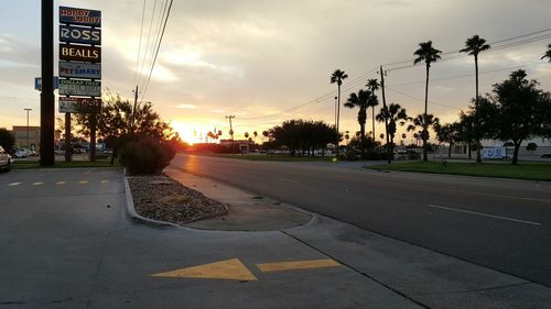 Empty road by coconut palm trees against sky during sunset