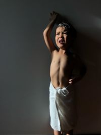 Shirtless boy wrapped in towel standing against gray wall