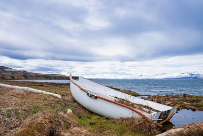 View of white boat moored on grassy field by seascape against cloudy sky