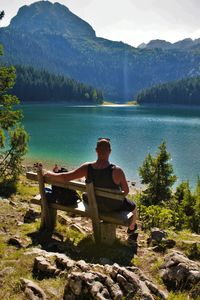 Rear view of young man looking at lake while sitting on bench during sunny day