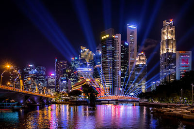Illuminated modern buildings by river against sky at night