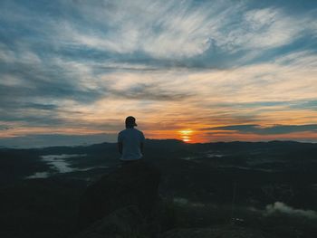 Rear view of man sitting on rock against landscape during sunset