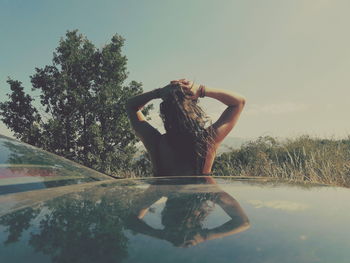 Rear view of woman with hands behind head standing by car against sky